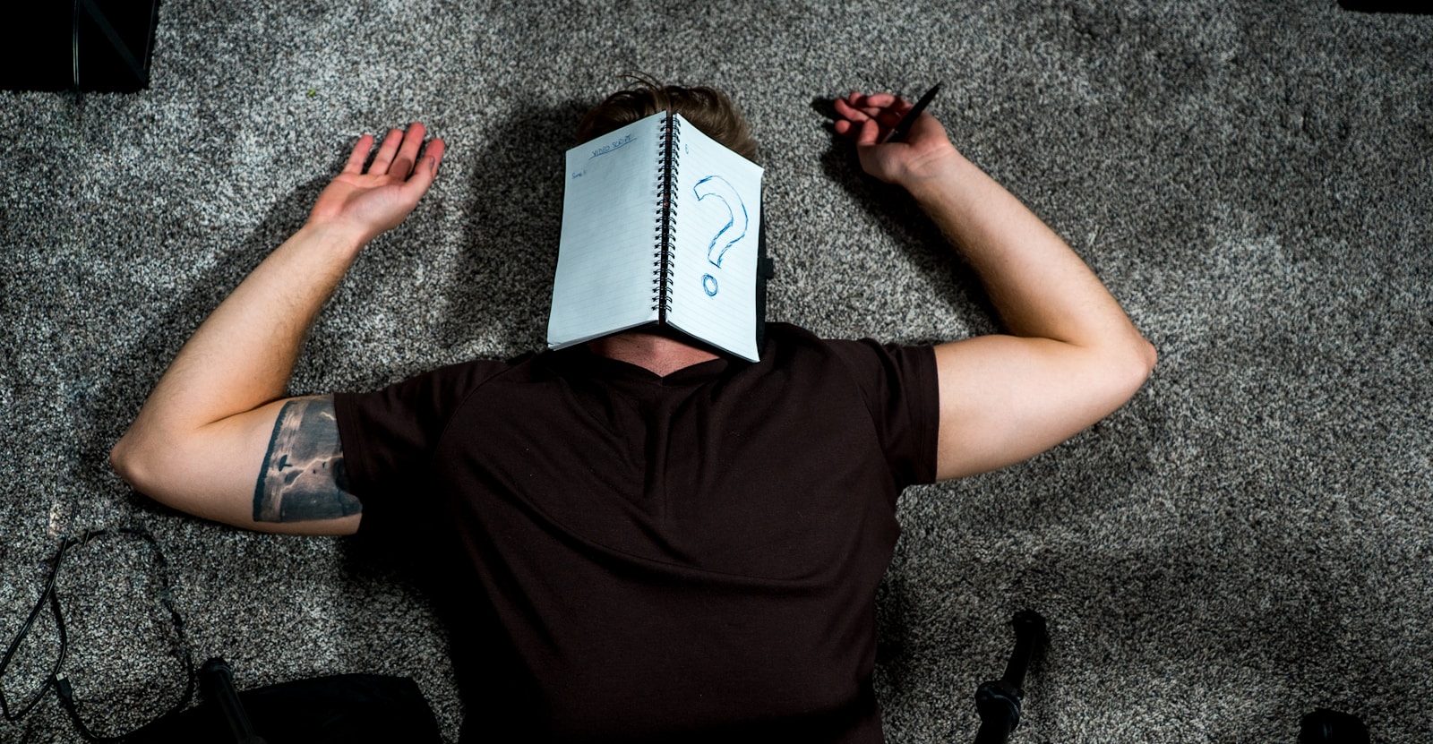 Person liegt auf dem Boden, wirkt erschöpft, geöffnetes Schreibheft mit Fragezeichen auf dem Gesicht drückt ihre Ratlosigkeit beim Schreiben vom Schlussteil der Arbeit aus