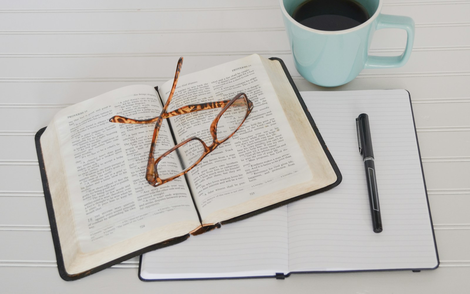 Ein theoretischer Rahmen basiert auf der Literaturrecherche, symbolisiert hier durch eine Brille auf einem aufgeschlagenen Buch, darunter ein aufgeschlagenes Notizbuch, darauf ein schwarzer Stift, daneben eine Tasse